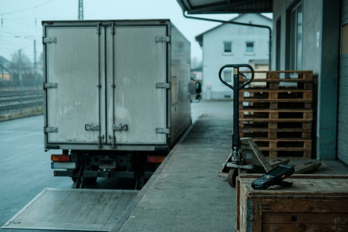 Box truck at a depot loading ramp beside pallets and a pallet truck